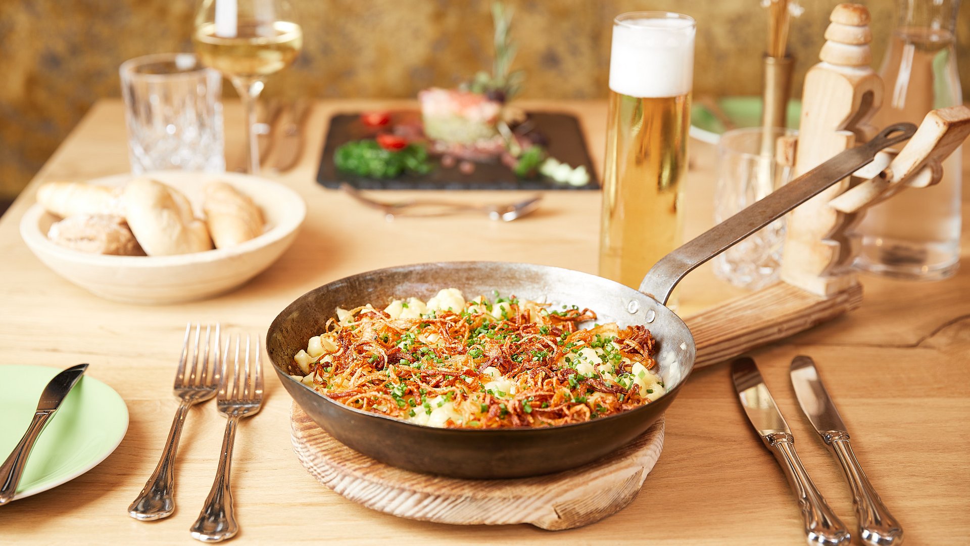 Spezialpfanne mit Kässpätzle und Röstzwiebeln auf Holztisch, dazu Bier und Brot