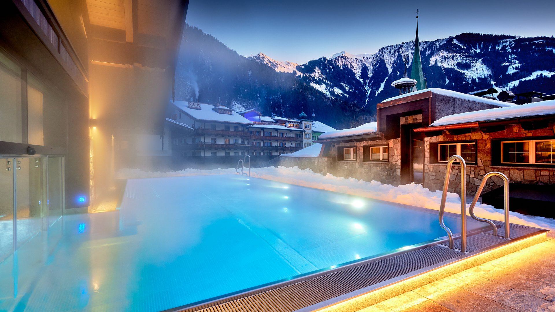 Beheizter Außenpool bei Schnee mit Bergkulisse und beleuchteten Gebäuden bei Abenddämmerung